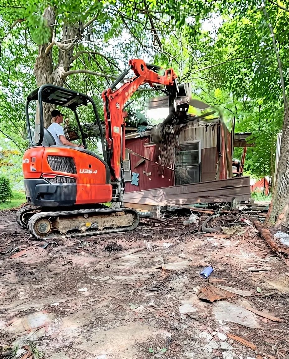 Excavator demolishing a small red structure in a wooded lot